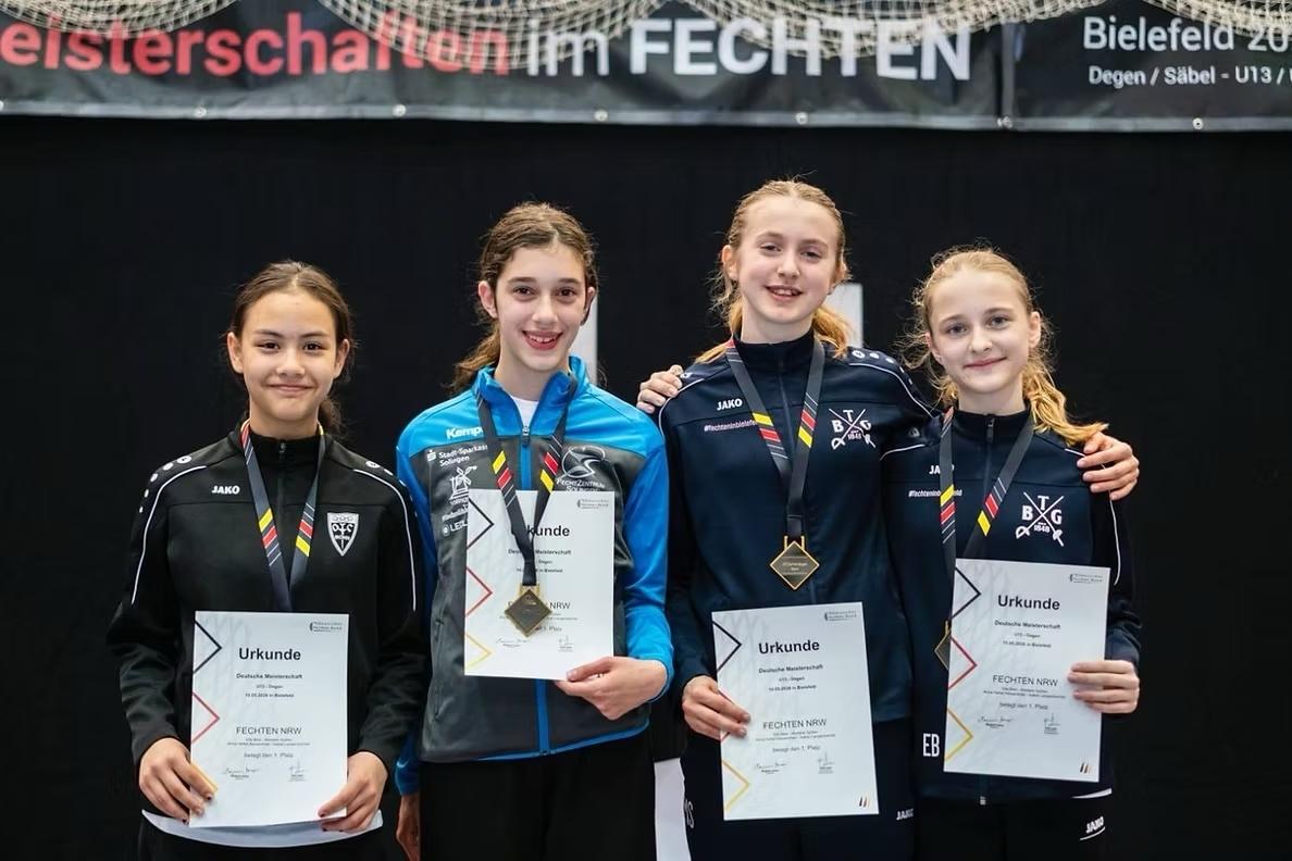 Vier fechtende Mädchen halten ihre Urkunden in der Hand, lächelnd, mit Medaillen um den Hals, im Hintergrund ein Banner.