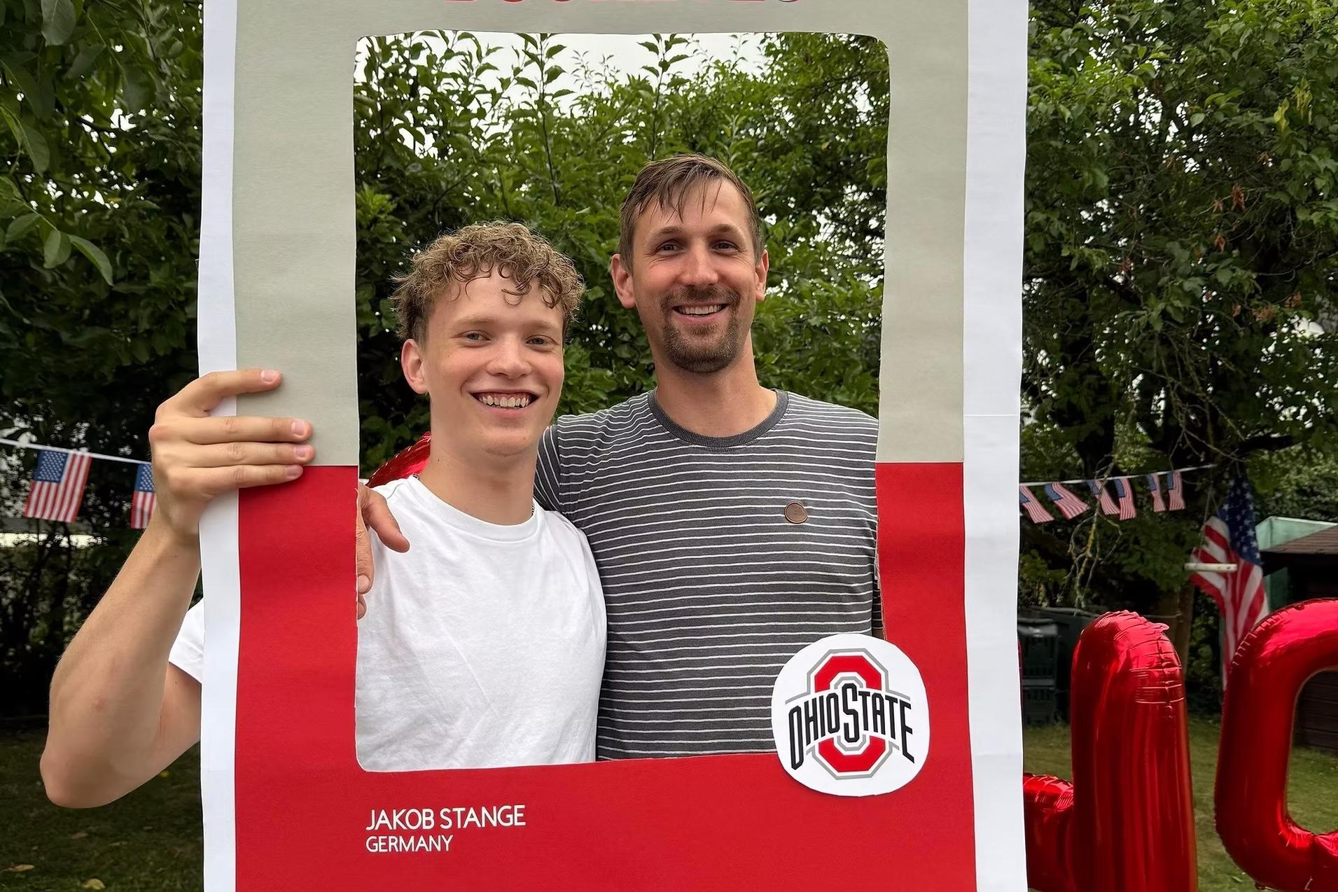 Zwei Männer posieren lächelnd vor einem großen, rahmenartigen Schild mit der Aufschrift „BUCKEYES“.