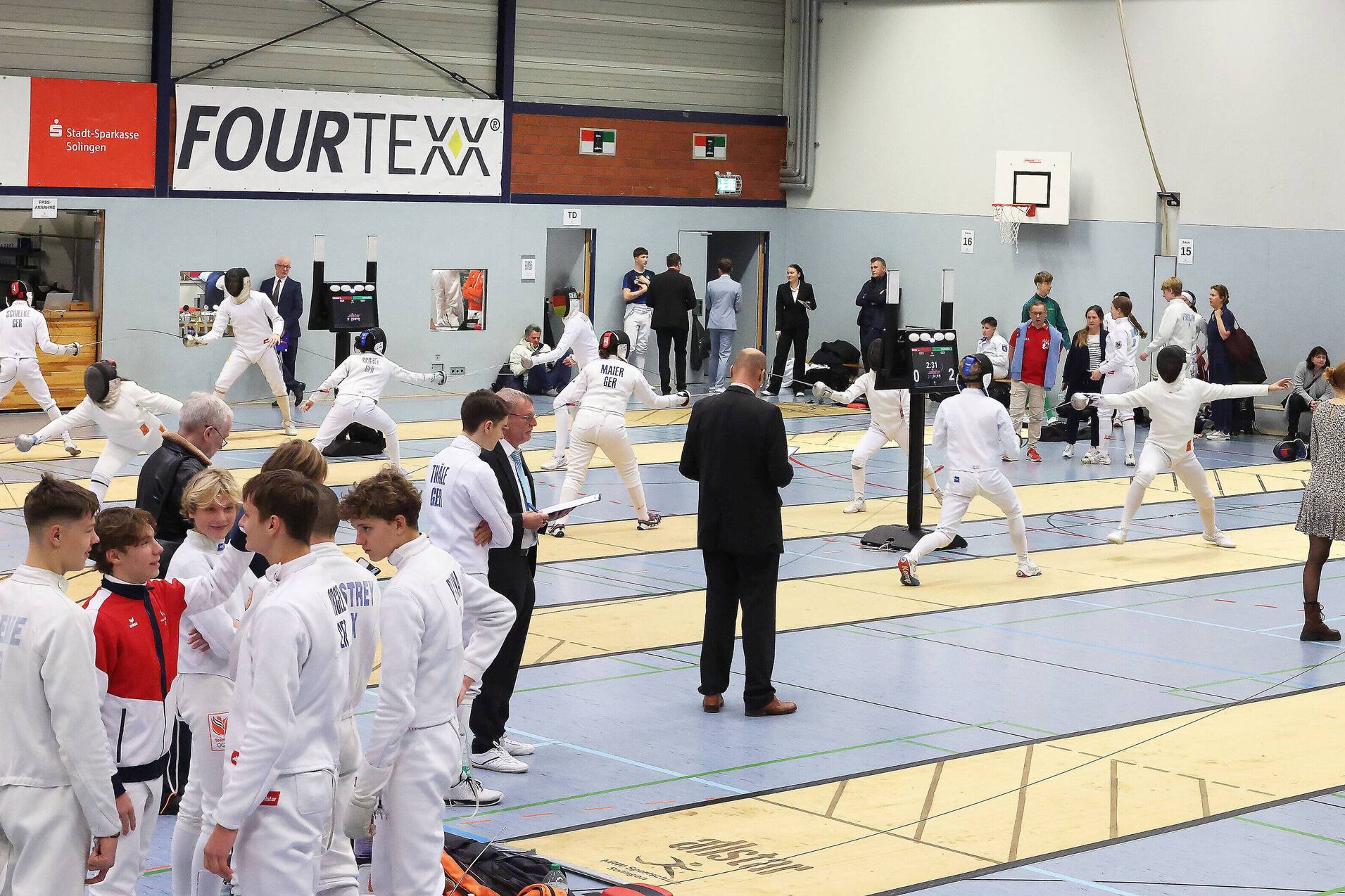 Fencers in white uniforms train auf mehreren Matten in einer Sporthalle, Zuschauer beobachten die Aktivitäten im Hintergrund.