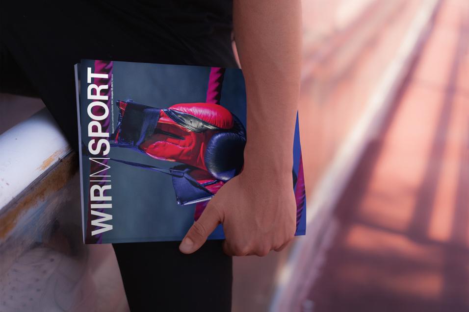 Nahaufnahme einer Hand, die ein Magazin mit dem Titel „WIR IM SPORT“ hält, während sie auf einer Sportbahn sitzt.